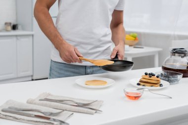 Cropped view of man holding pancake and frying pan near coffee and honey in kitchen  clipart