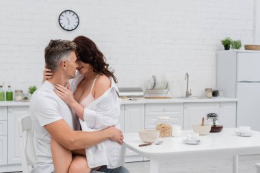 Sexy woman in bra and shirt kissing husband near breakfast in kitchen  clipart