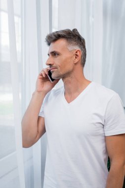 Beyaz tişörtlü adam evde akıllı telefondan konuşuyor. 