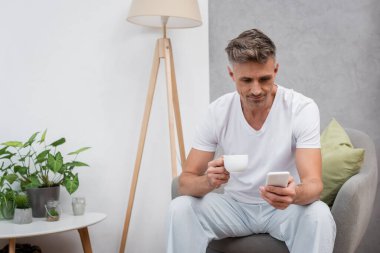 Elinde kahve tutan ve evdeki koltukta akıllı telefon kullanan bir adam. 