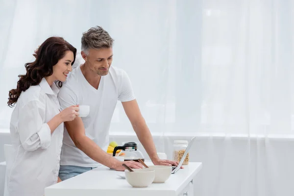 在厨房用笔记本电脑在咖啡和麦片早餐旁边微笑的夫妻 — 图库照片