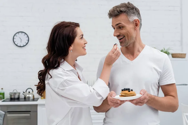 Leende Kvinna Håller Färska Blåbär Nära Make Med Pannkakor Köket — Stockfoto