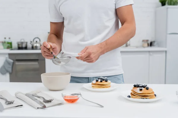 Cropped view of man pouring powdered sugar in sieve near honey and pancakes in kitchen