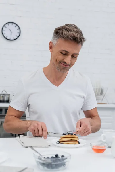 Positieve Man Snijden Pannenkoeken Buurt Van Bosbessen Honing Keuken — Stockfoto