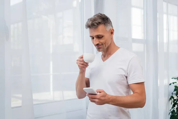 Hombre Camiseta Blanca Usando Teléfono Celular Sosteniendo Taza Café Casa —  Fotos de Stock