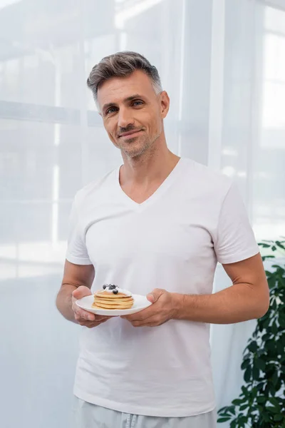Smiling man in pajamas holding pancakes and looking at camera at home