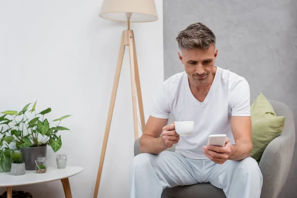 男人在家里拿着一杯咖啡 在扶手椅上使用智能手机 — 图库照片