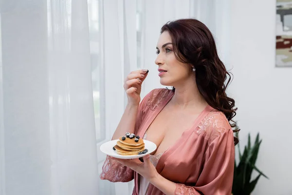 Brunette woman in silk robe holding blueberry and pancakes at home