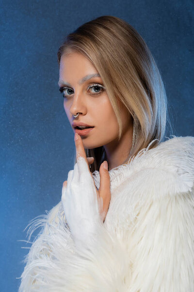 blonde woman with winter makeup and frozen eyebrows touching lip on dark blue 