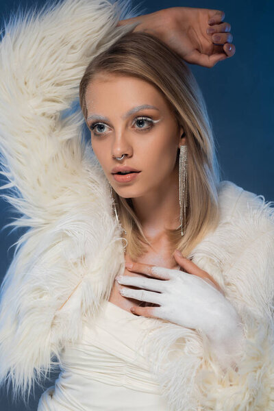 pierced young woman with winter makeup posing in white jacket with feathers on dark blue 