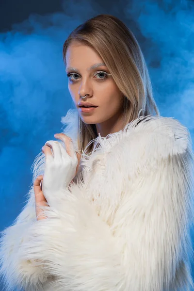 冬の化粧をしたピアスの女性は煙と青い上の羽を持つフェイクファーのジャケットにポーズ — ストック写真