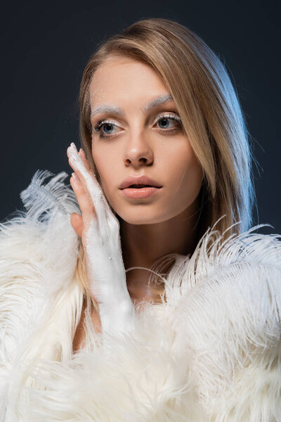 blonde young woman with winter makeup posing in white faux fur jacket and looking away isolated on blue