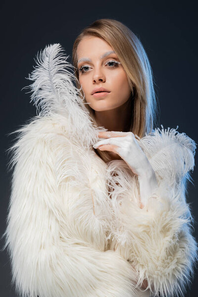 pretty young woman in white faux fur jacket posing with feather isolated on blue