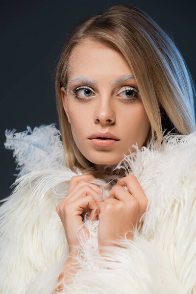 portrait of blonde young woman in faux fur jacket posing with white feathers isolated on blue