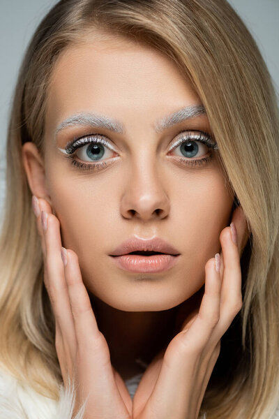 close up view of young model with winter makeup and frozen white eyebrows touching face isolated on grey