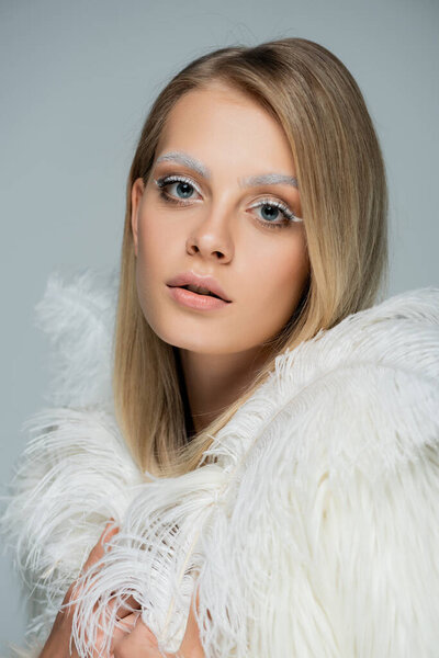 portrait of pretty woman with winter makeup and white eyebrows looking at camera near white feather isolated on grey