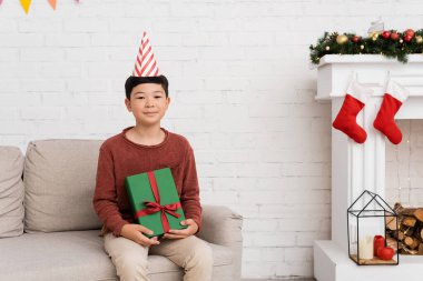 Parti şapkalı ergen Asyalı çocuk evdeki Noel dekorunun yanındaki koltukta hediyesini tutuyor. 