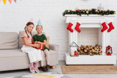 Parti şapkalı gülümseyen arkadaşlar evdeki Noel dekorunun yanındaki kanepede hediyelerini tutuyorlar. 