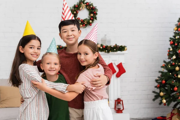 誕生日や家での新年のお祝い中に抱きしめるパーティーキャップで多民族の子供たちを笑顔 — ストック写真