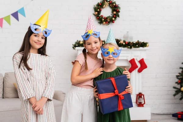 Smiling Girls Party Caps Masks Hugging Friend Present Christmas Decor — Stock Photo, Image