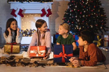 Gülümseyen çoklu etnik çocuklar Noel hediyeleri ve şöminenin yanında konuşuyorlar. 
