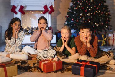 Hediyelerin yakınındaki kameraya bakan korkmuş çoklu etnik çocuklar ve akşam evde bulanık Noel ağacı. 