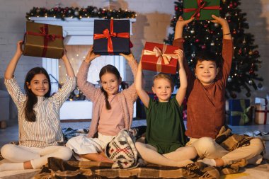 Çok ırklı çocuklar ellerinde hediyelerle şöminenin yanındaki battaniyeye oturmuş evde Noel süsü veriyorlar. 