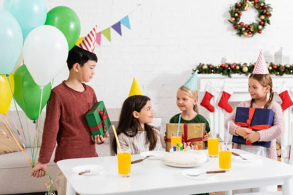 在家里 戴着派对帽的多种族孩子在朋友家附近拿着礼物和气球 还拿着生日蛋糕 — 图库照片