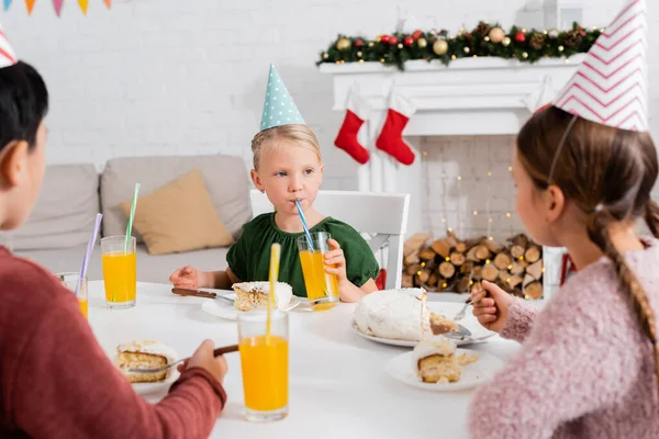 戴着派对帽的孩子在模糊的朋友身边喝橙汁 在家里吃生日蛋糕 — 图库照片