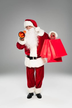 Noel Baba kostümlü, gözlüklü, hoparlörlü ve gri arka planda alışveriş çantalı. 