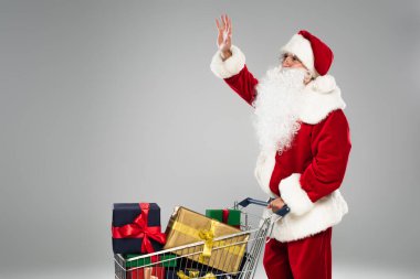 Noel Baba şapkalı, alışveriş arabasının yanında el sallıyor, hediyeler gri renkte. 