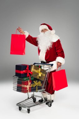 Noel Baba alışveriş çantalarını tutuyor ve gri arka planda hediyelerin yanında kameraya bakıyor. 