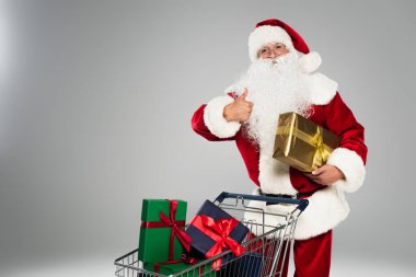 Noel Baba jest yapıyor ve gri üzerinde izole edilmiş alışveriş arabasının yanında duruyor. 