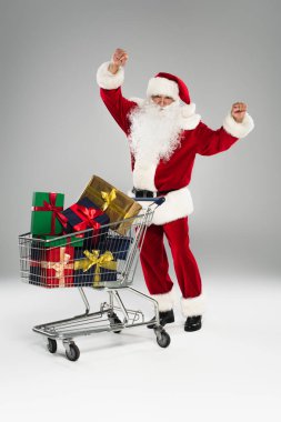 Heyecanlı Noel Baba, gri arka planda hediyelerle alışveriş arabasının yanında duruyor. 