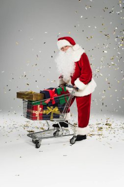 Noel Baba elinde hediyelerle alışveriş arabasıyla gri arka planda konfeti altında duruyor. 