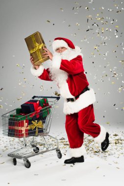 Noel Baba alışveriş arabasının yanında duruyor ve gri arka planda konfeti var. 