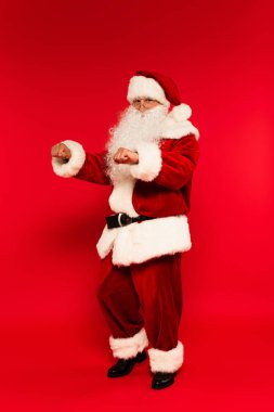 Sakallı Noel Baba kırmızı arka planda dans ederken kameraya bakıyor. 