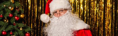 Bearded santa claus looking at camera near christmas tree and tinsel, banner  clipart
