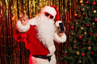 Güneş gözlüklü Noel Baba boş ekranlı akıllı telefonu tutuyor ve Noel ağacı ve süs eşyalarının yanında parmakla işaret ediyor. 