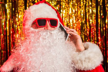 Bearded santa claus in sunglasses talking on smartphone near blurred tinsel  clipart