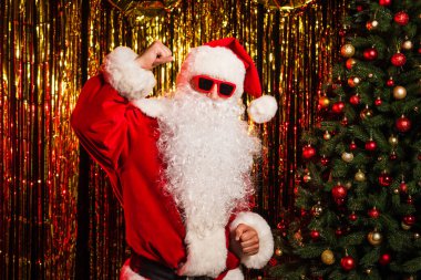 Noel Baba güneş gözlüğü ve kostüm takmış Noel ağacı ve süs eşyalarının yanında dans ediyor. 