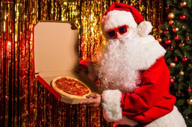 Güneş gözlüklü Noel Baba, Noel ağacı ve süs eşyalarının yanında hazır pizza tutuyor. 