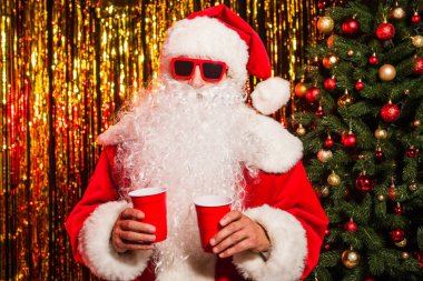 Santa claus in sunglasses and hat holding plastic cups near christmas tree and tinsel  clipart