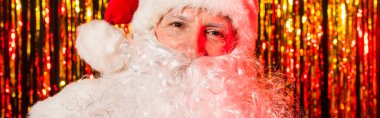 Portrait of santa claus in red hat looking at camera near blurred tinsel, banner  clipart