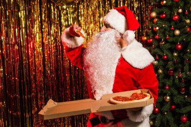 Noel Baba kostüm içinde, Noel ağacı ve süs eşyalarının yanında nefis bir pizza tutuyor. 