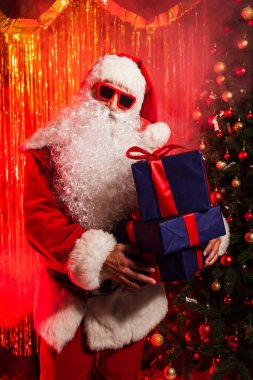 Güneş gözlüklü Noel Baba Noel ağacının yanında hediyeler ve bulanık süslemeler taşıyor. 