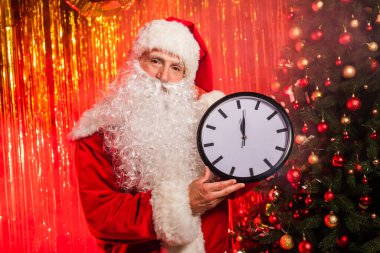 Noel Baba kostüm içinde Noel ağacı ve süs eşyalarının yanında saat tutuyor. 