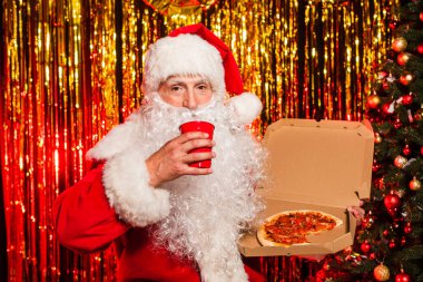 Santa claus in costume holding plastic cup and takeaway pizza near christmas tree and tinsel  clipart