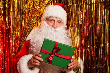 Santa claus in costume holding present and looking at camera near tinsel  clipart