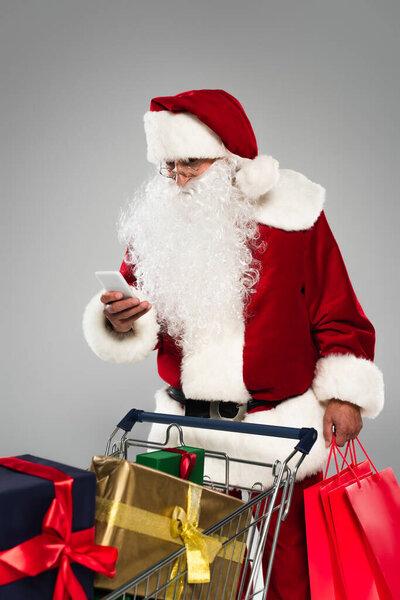 Santa claus using mobile phone and holding shopping bags near gifts in cart isolated on grey 